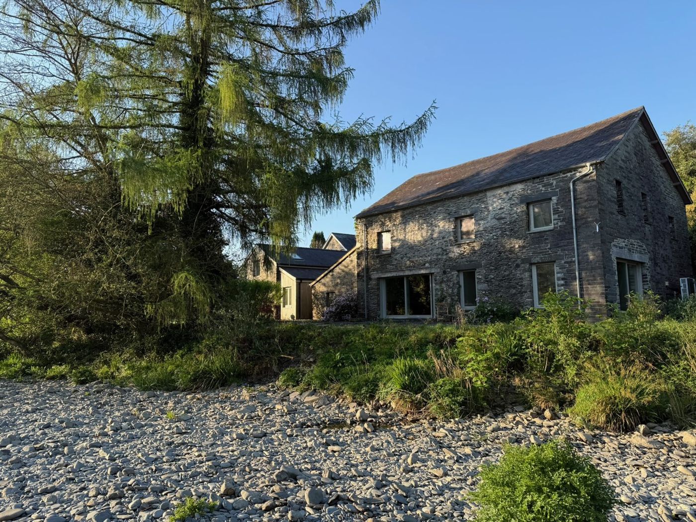 The Mill: a stone barn with a slate roof and full-height windows, seen from the river’s stony bank