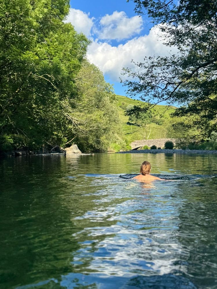 A swimmer in the river with the old stone bridge and green hillside beyond